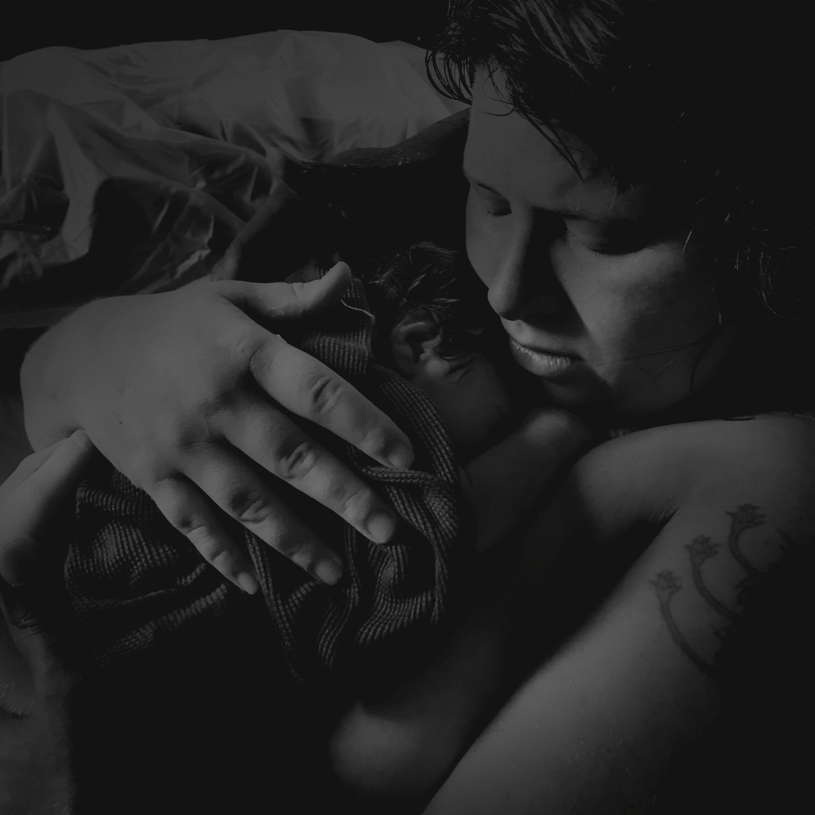 Mother's eyes closed, her newborn against her cheek, the father's hand at her shoulder — two minutes old